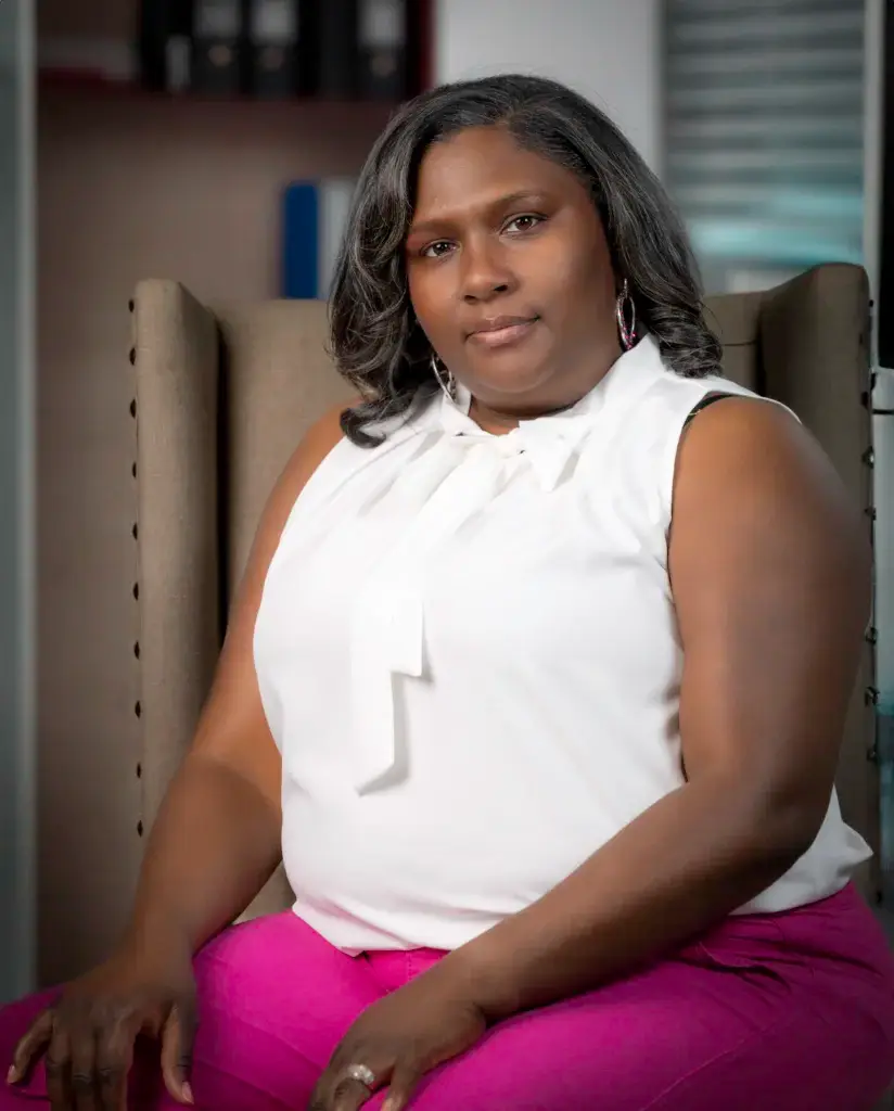 Torrie Powell, CLINICAL PSYCHOTHERAPIST with shoulder-length dark hair, wearing a sleeveless white blouse and bright pink pants, is seated in a beige upholstered chair in an office setting. She is looking directly at the camera with a calm and composed expression.